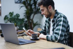 Young man at his dining room table, smiling as he looks at a laptop and his smartphone