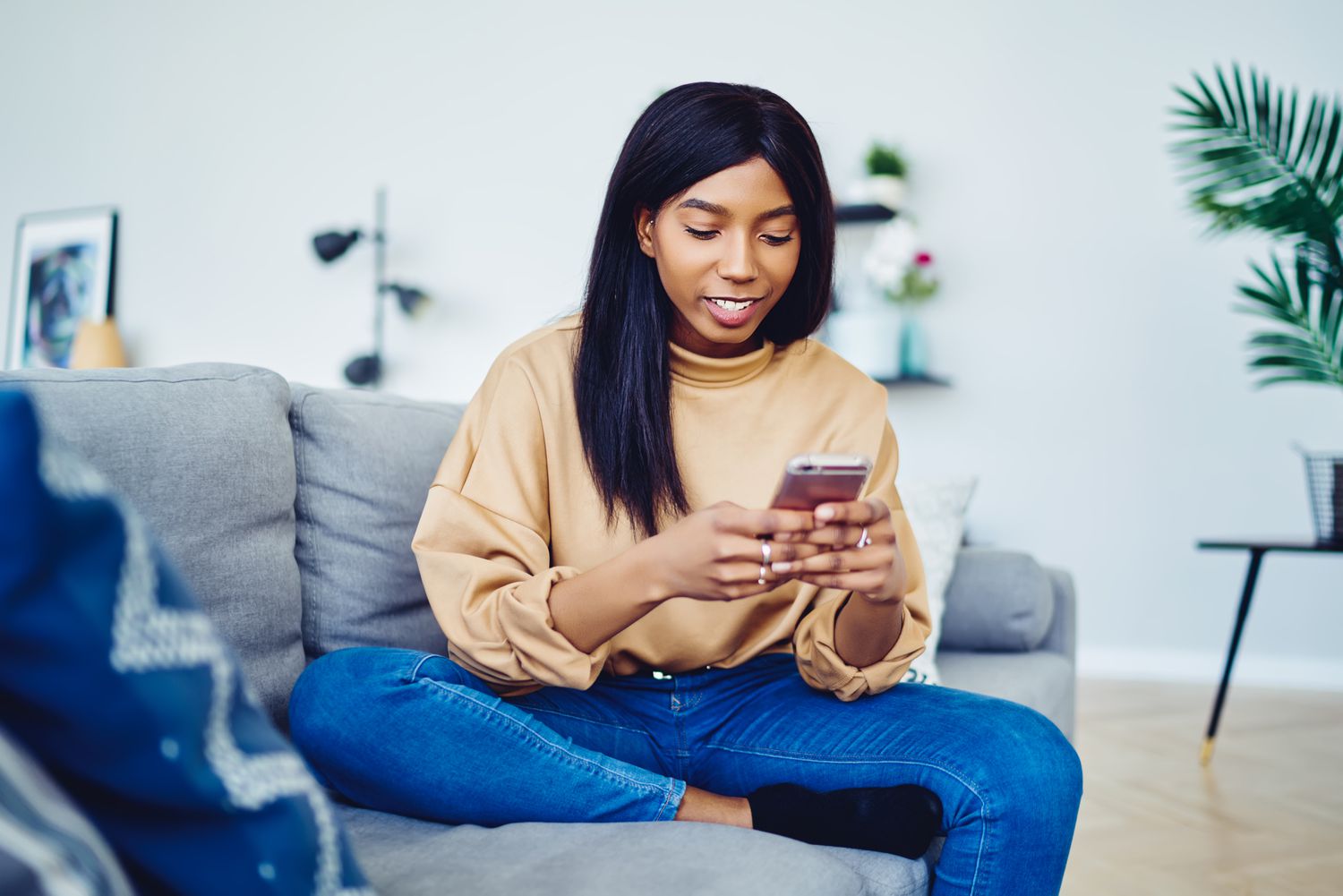 Young woman sitting on a living room couch and looking with a smile at something on her smartphone