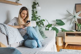 Young woman sitting on her couch and looking happily at something on her smartphone