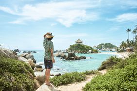 Person standing on a rocky beach looking towards a gazebo-like structure on a small island near a tropical coastline