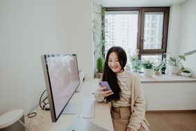 Woman in her home office with a large monitor on the desk, smiles while looking at her phone.