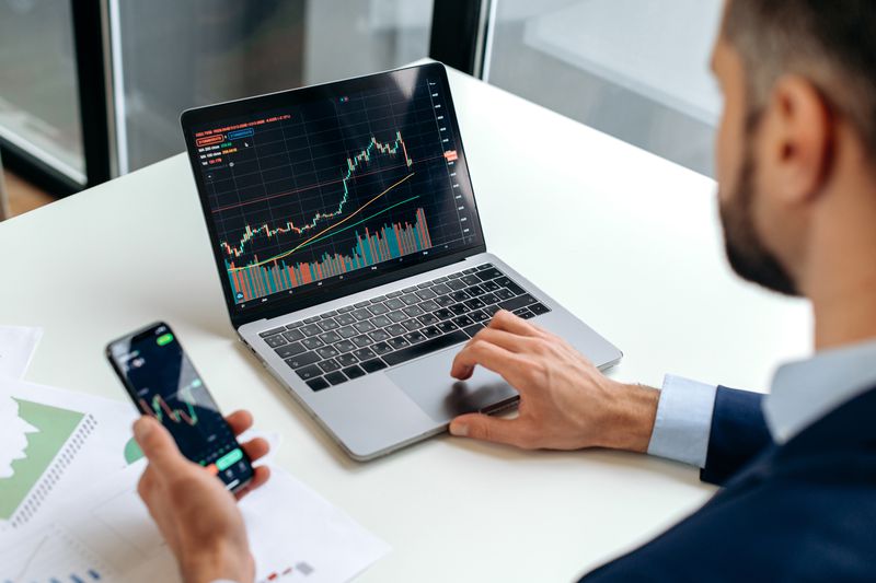 Trader checking market index on laptop while making trading decision on the phone.
