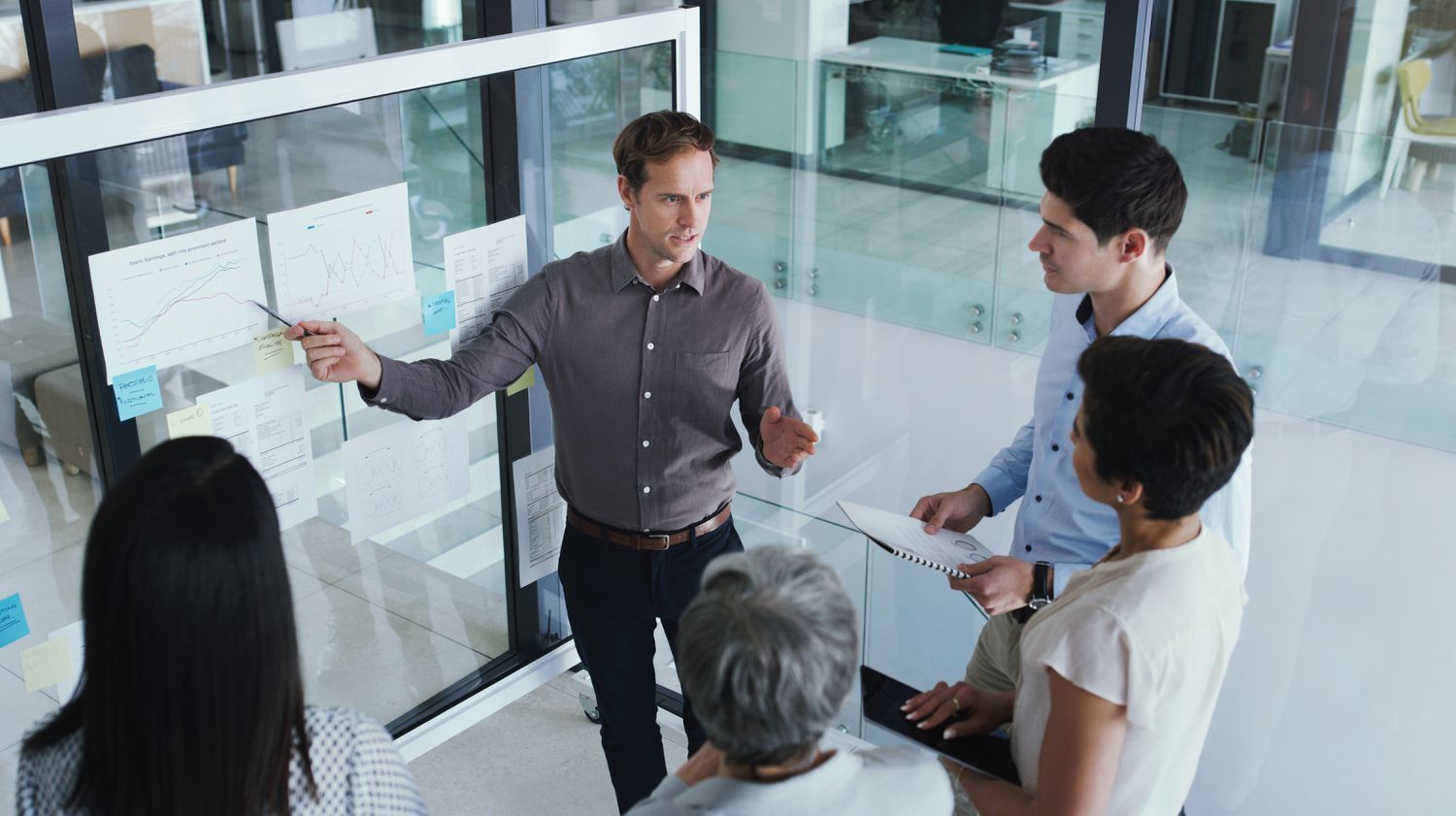 A group of office workers standing in a semi circle as one man points at a roller white board with papers taped to it, shot from above. 