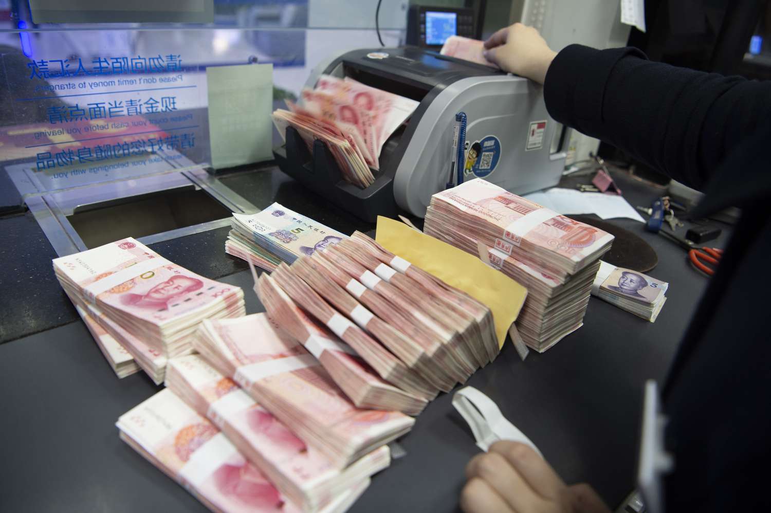A staff member of a bank standing behind glass and feeding stacks of banknotes into a cash counter.
