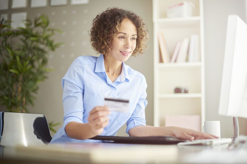 A young woman sits at her desk looking at a computer, holding a bank card in her hand.