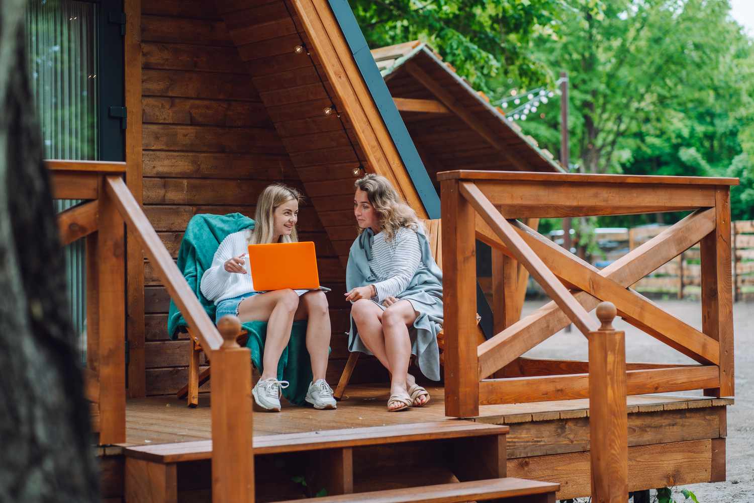 Two women sitting on chairs outside of a cabin, with one holding a laptop.