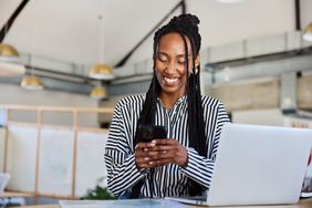 Young person smiling and texting on a smartphone while working on a laptop at a desk in an office.