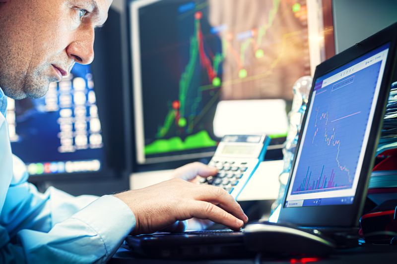 Profile view of a stockbroker calculating after-cost profit of possible trades while they focus on a screen showing stock data and use a keyboard.