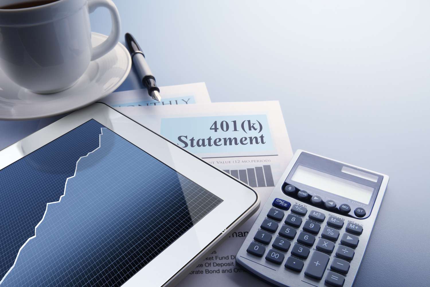 A digital tablet with a stock market chart on its screen rests on top of a 401(k) statement that sits on a desktop. A fountain pen, a calculator, and cup of coffee also frame the scene.