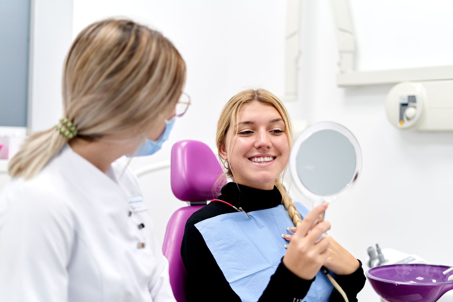 A woman sits in a dentist chair next to a female dentist looking in a mirror at her teeth