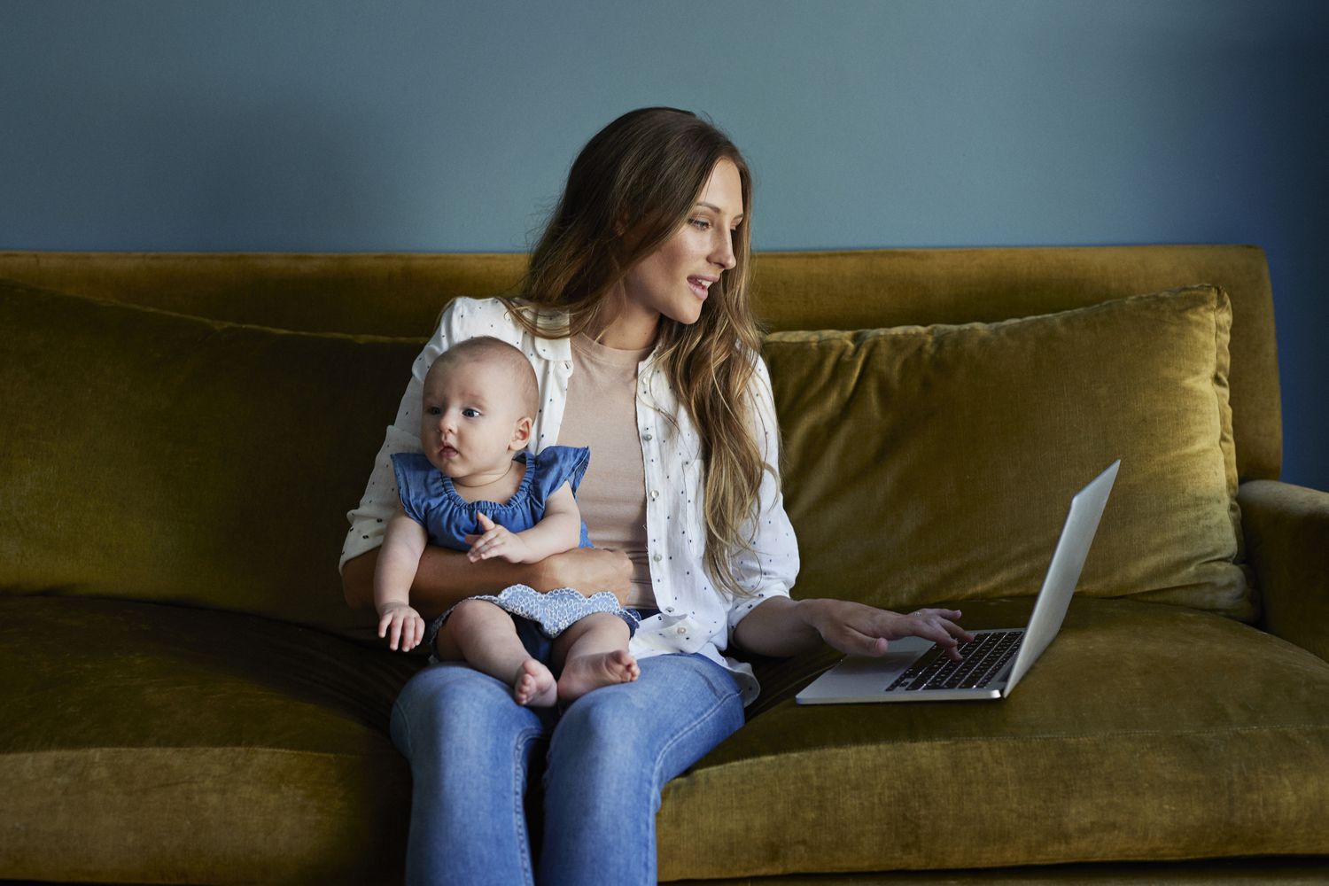 A woman holds a baby on her lap while sitting on a couch and working on a laptop