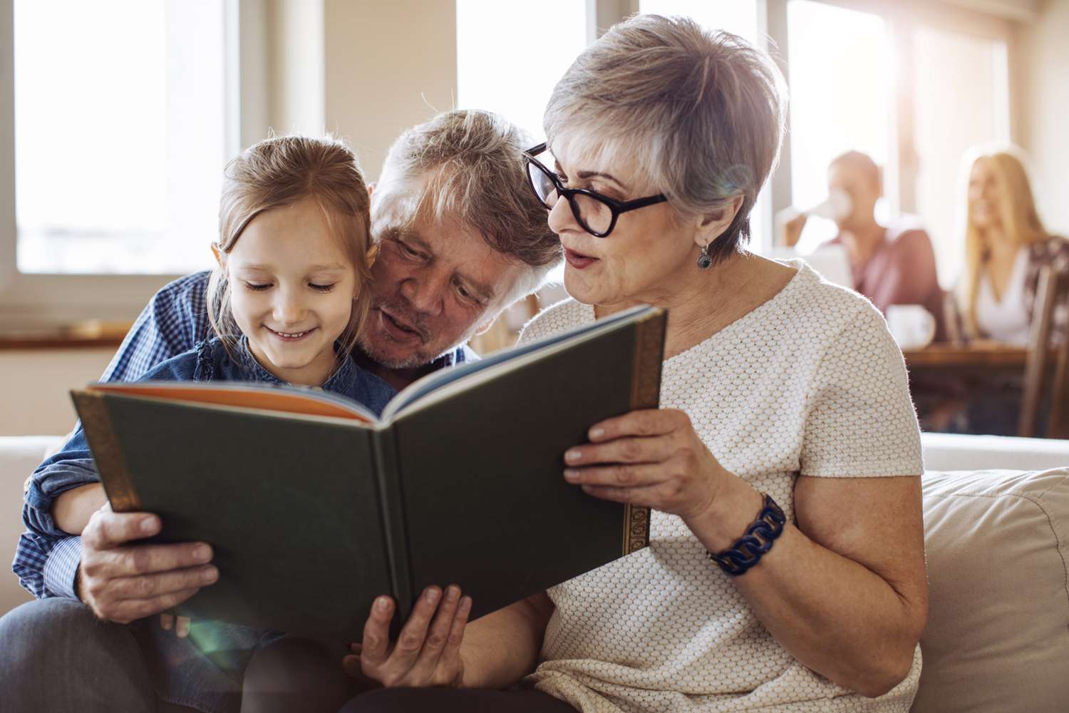 Grandparents reading fairy tale to granddaughter at home with the parents drinking coffee behind them.