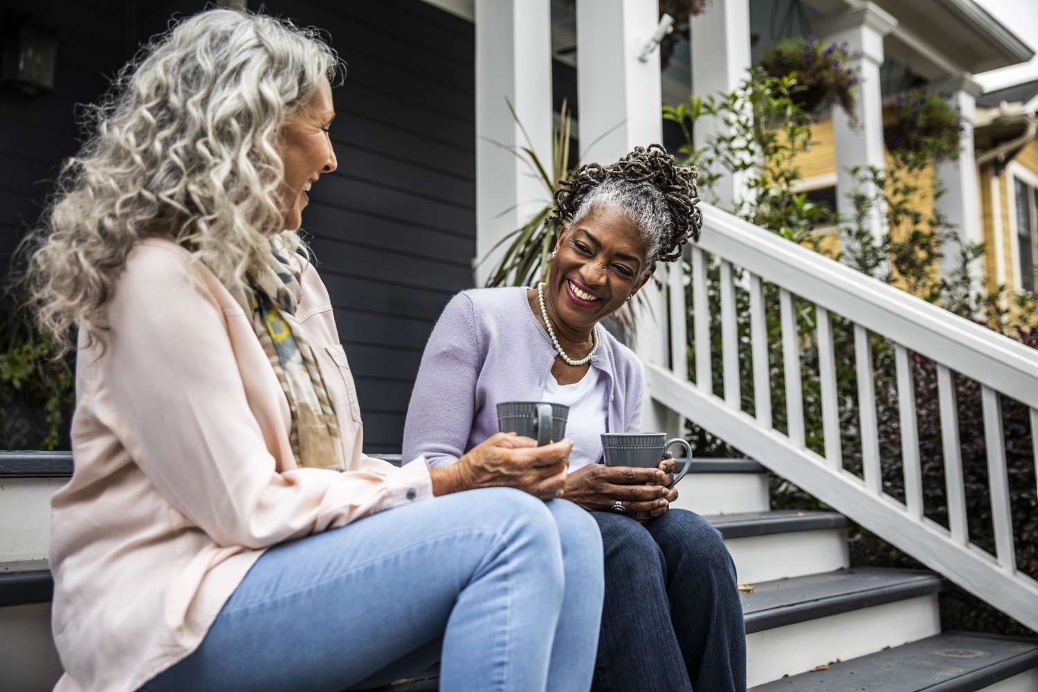 Two older woman sitting on the porch of a suburban home laughing and drinking coffee.