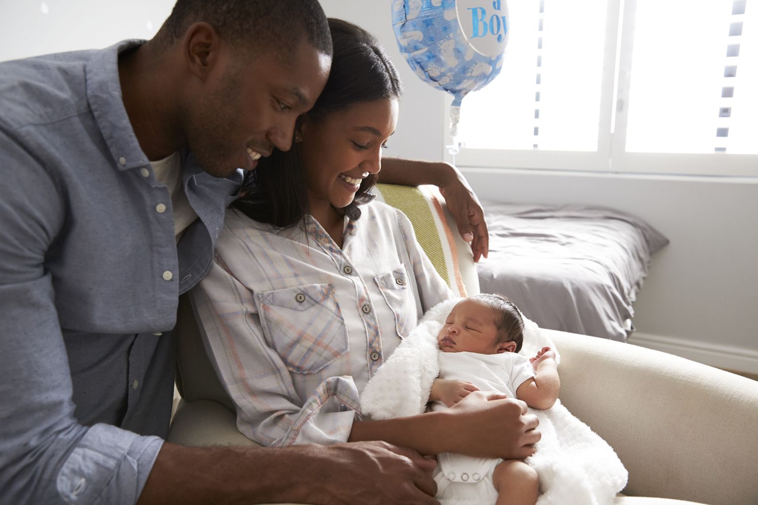 Two parents hold a newborn baby in a chair at the hospital with a balloon in the background that says "Boy" on it