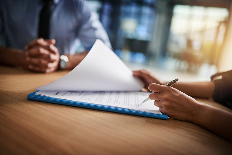 A person seated at a table looks through a document while another person waits.