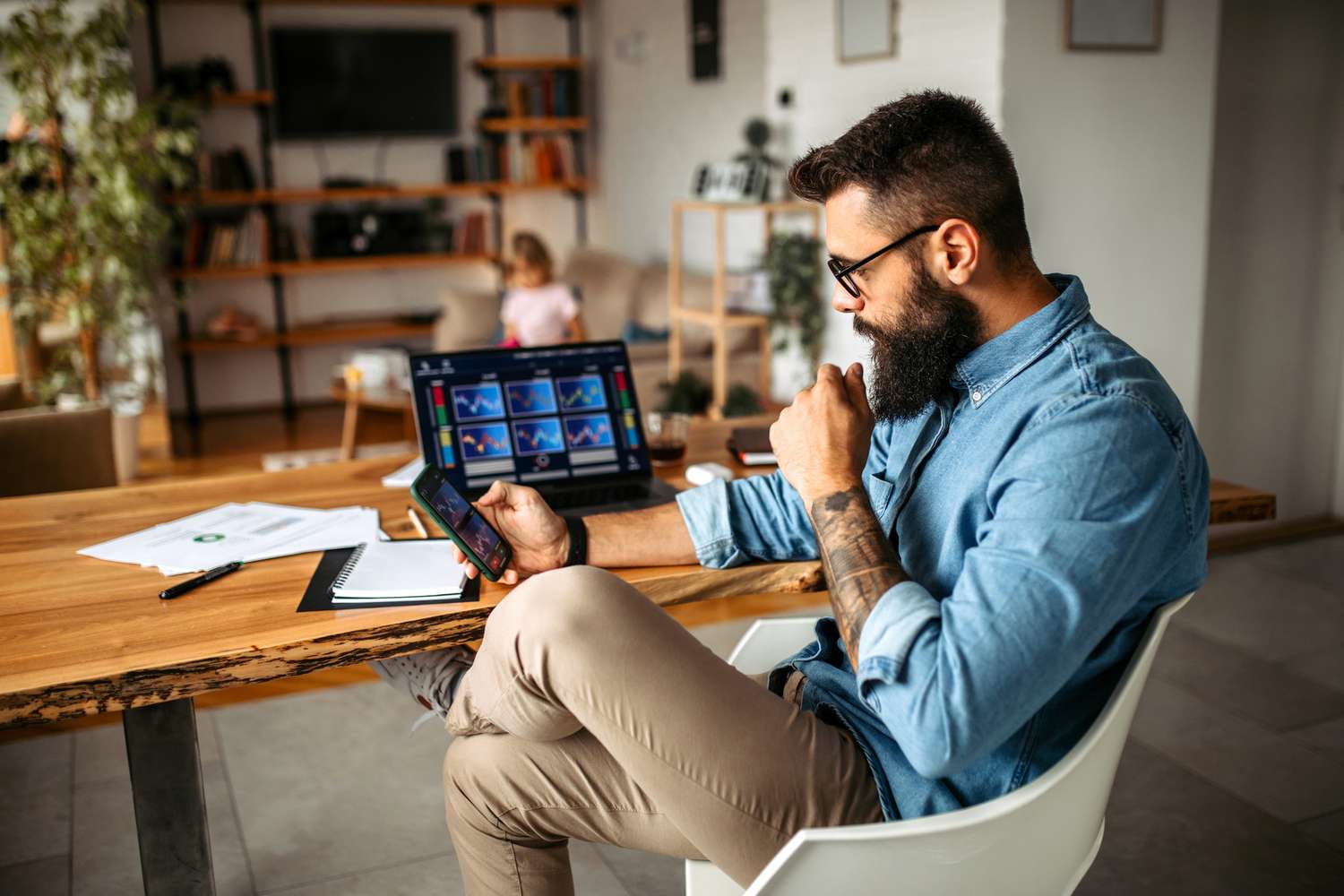Man with a beard sits at a kitchen table holding a smartphone in front of a laptop and both screens are showing stock charts. A child sits in the background.