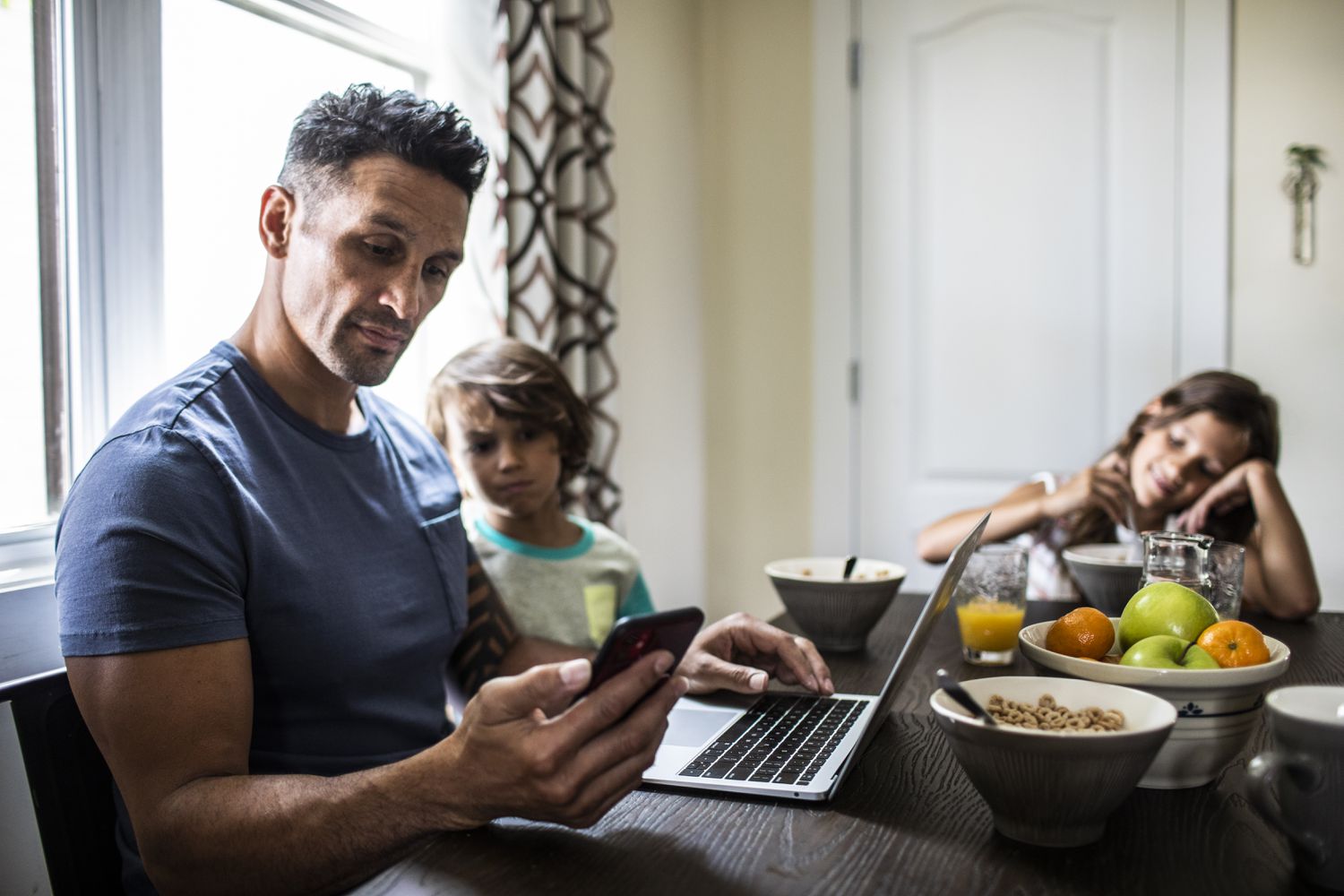 man looks at phone while on computer with kids at breakfast table