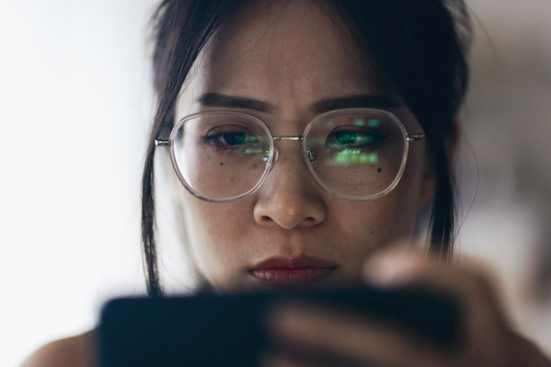 A woman holding a smartphone that is being reflected in her glasses with green charts and/or words