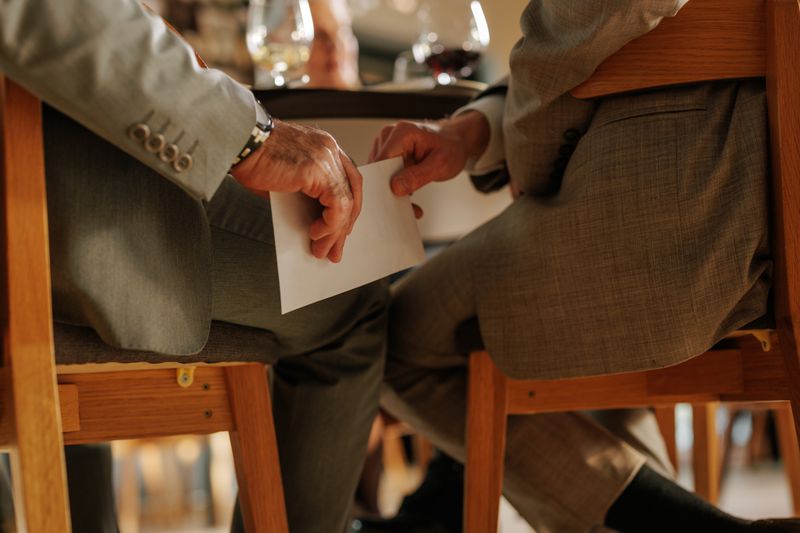 Two business executives clandestinely exchange an envelope of company secrets below a dinner table. 
