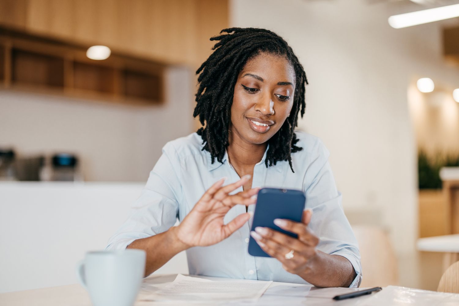A woman sits at a table with a mug, pen, papers, and a smartphone