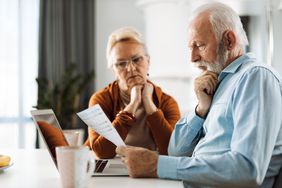 An older man and older woman sit at a table with a laptop and mug looking at a document