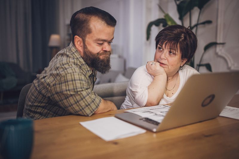 Worried couple sitting at a desk with an open laptop reviewing bills.