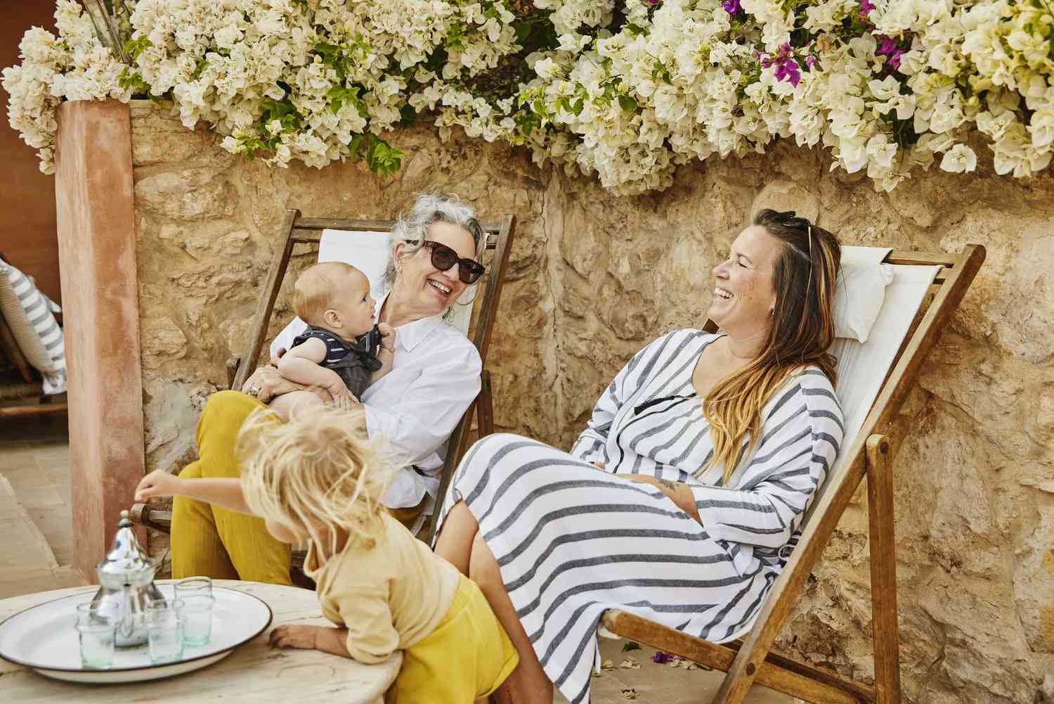 Wide shot of grandmother holding grandson in lap while laughing with daughter at tropical villa during multigenerational family vacation