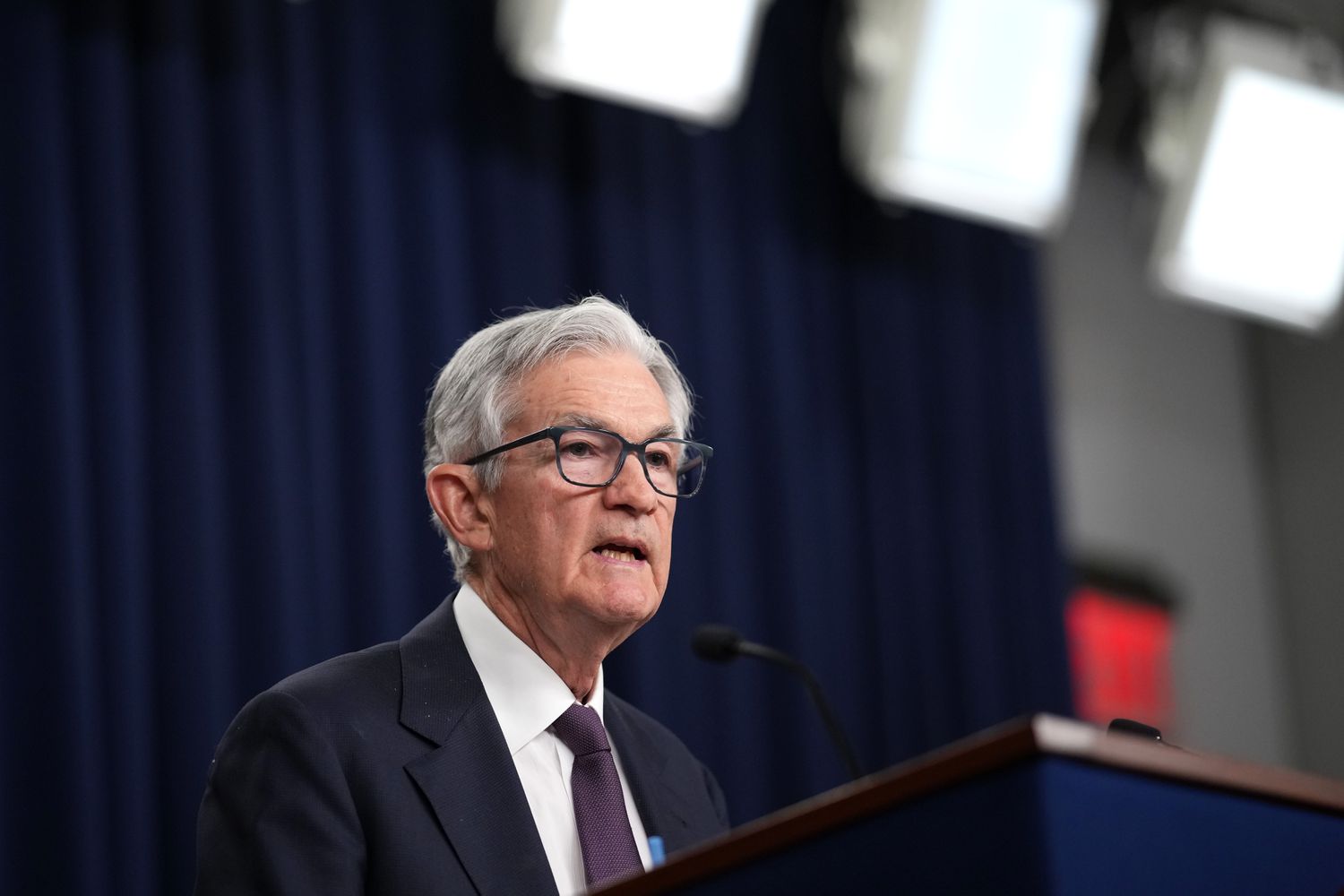 Jerome Powell stands in front of a podium, with curtains behind him during a press conference.