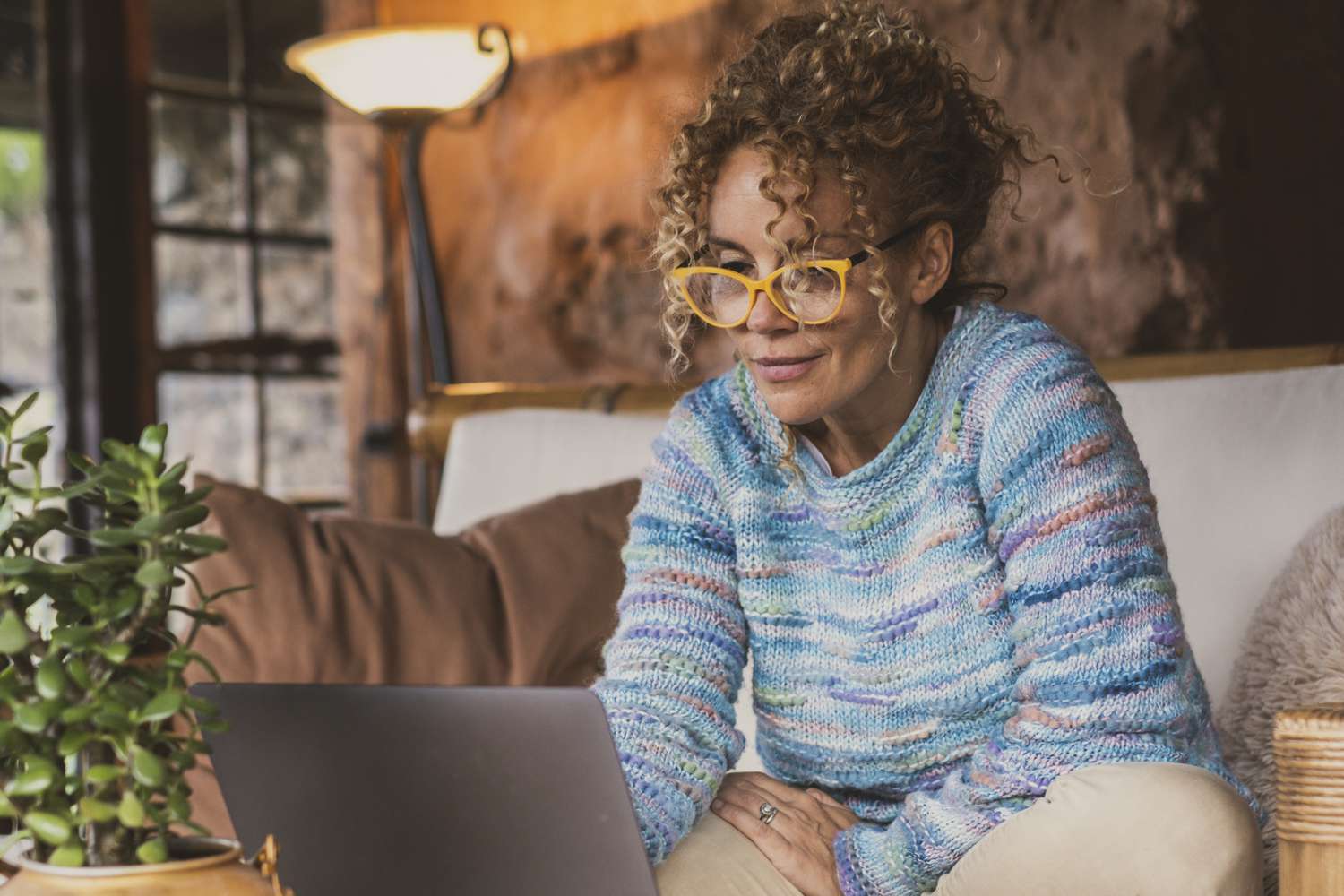 A woman with a blue sweater on sits on a couch wearing yellow glasses as she looks at a laptop on the coffee table