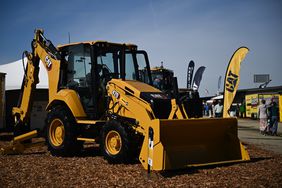 A yellow Caterpillar backhoe sits on a patch of mulch, with a banner bearing the company's black and yellow CAT logo to the right.