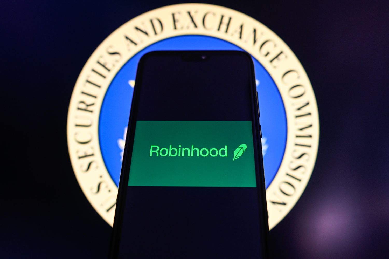 The green Robinhood logo is seen on a phone screen in front of the Securities and Exchange Commission seal in the background.