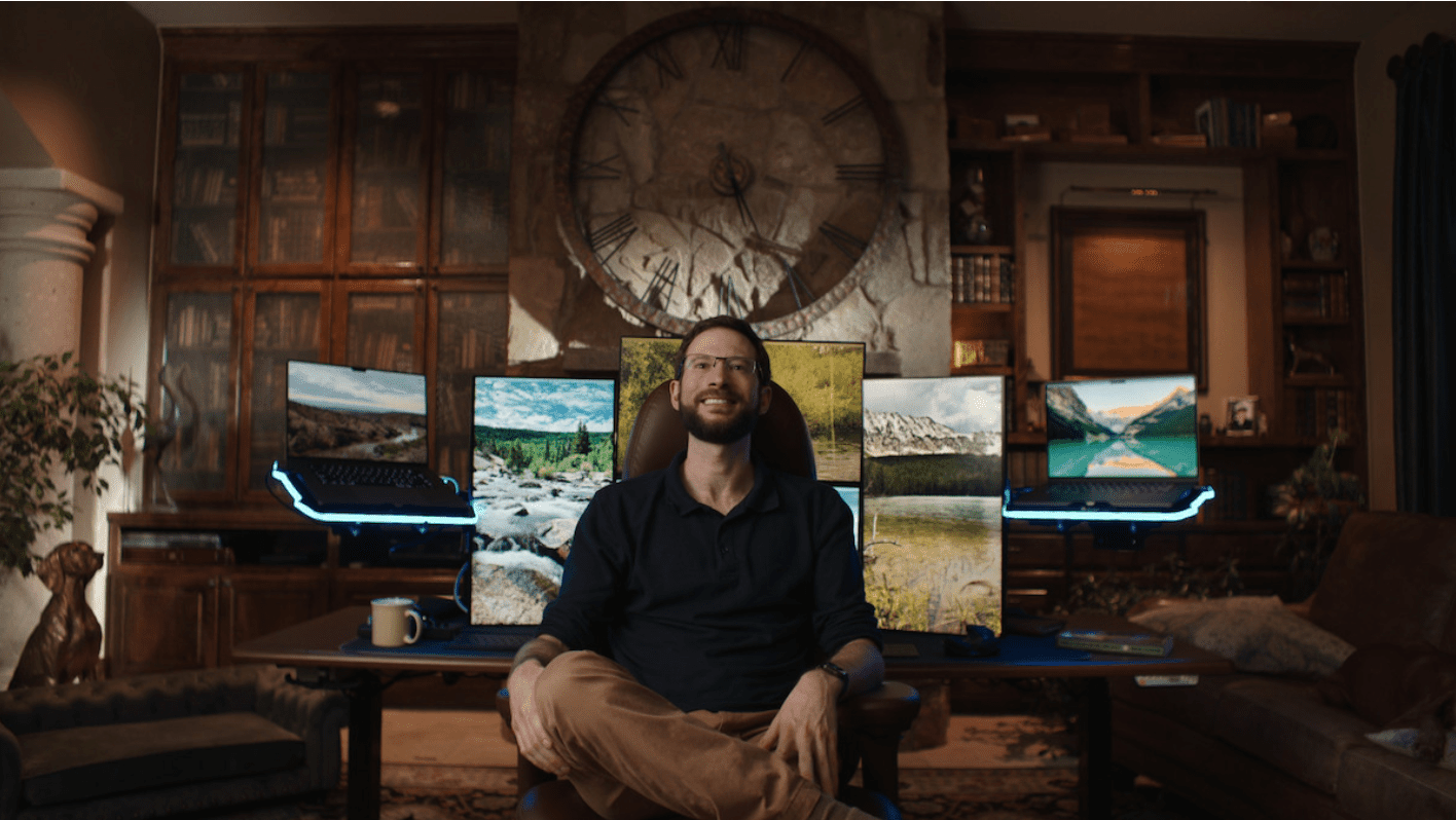 Person seated in front of a desk with multiple computer monitors in a room with a large wall clock