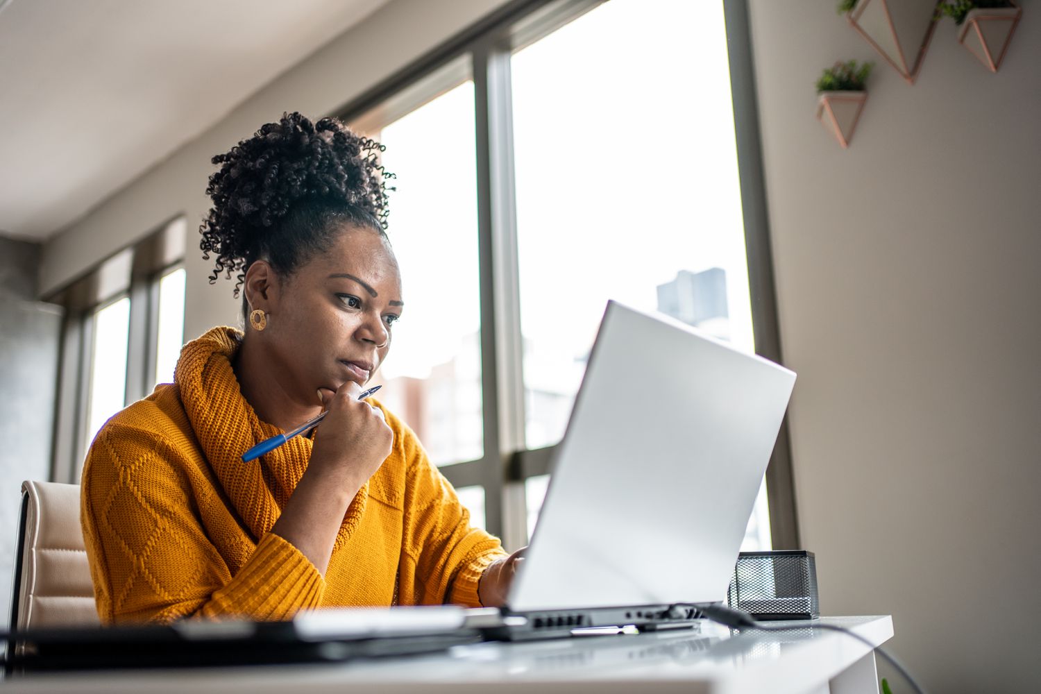 A woman wearing a yellow sweater sits at a desk at home looking at a laptop
