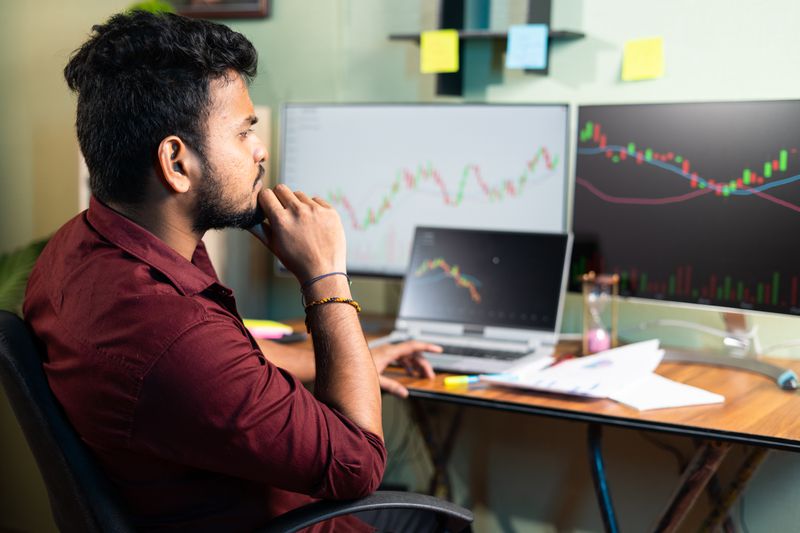 Stock trader sitting in the office while monitoring indexes of Indian stock market on 3 different screens.