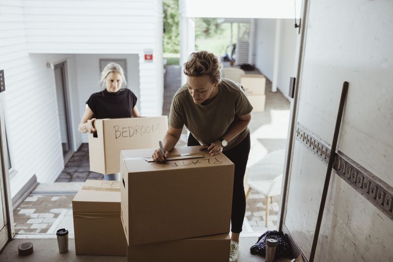 Person writing on a cardboard box inside a moving van while another holds a box outside