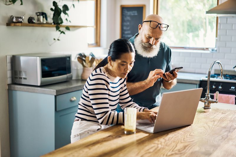 A woman works on a laptop at a kitchen counter while a man stands and uses a smartphone