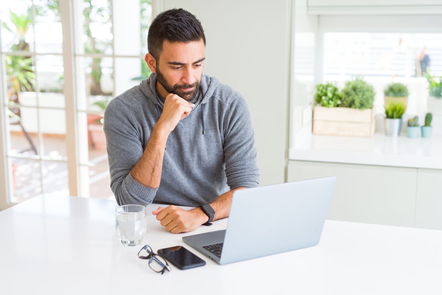 Man in his 30s sitting at his kitchen table and pondering something on his laptop