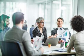 A company board meets around a conference table holding financial documents to discuss financing expansion through the sale of corporate bonds.