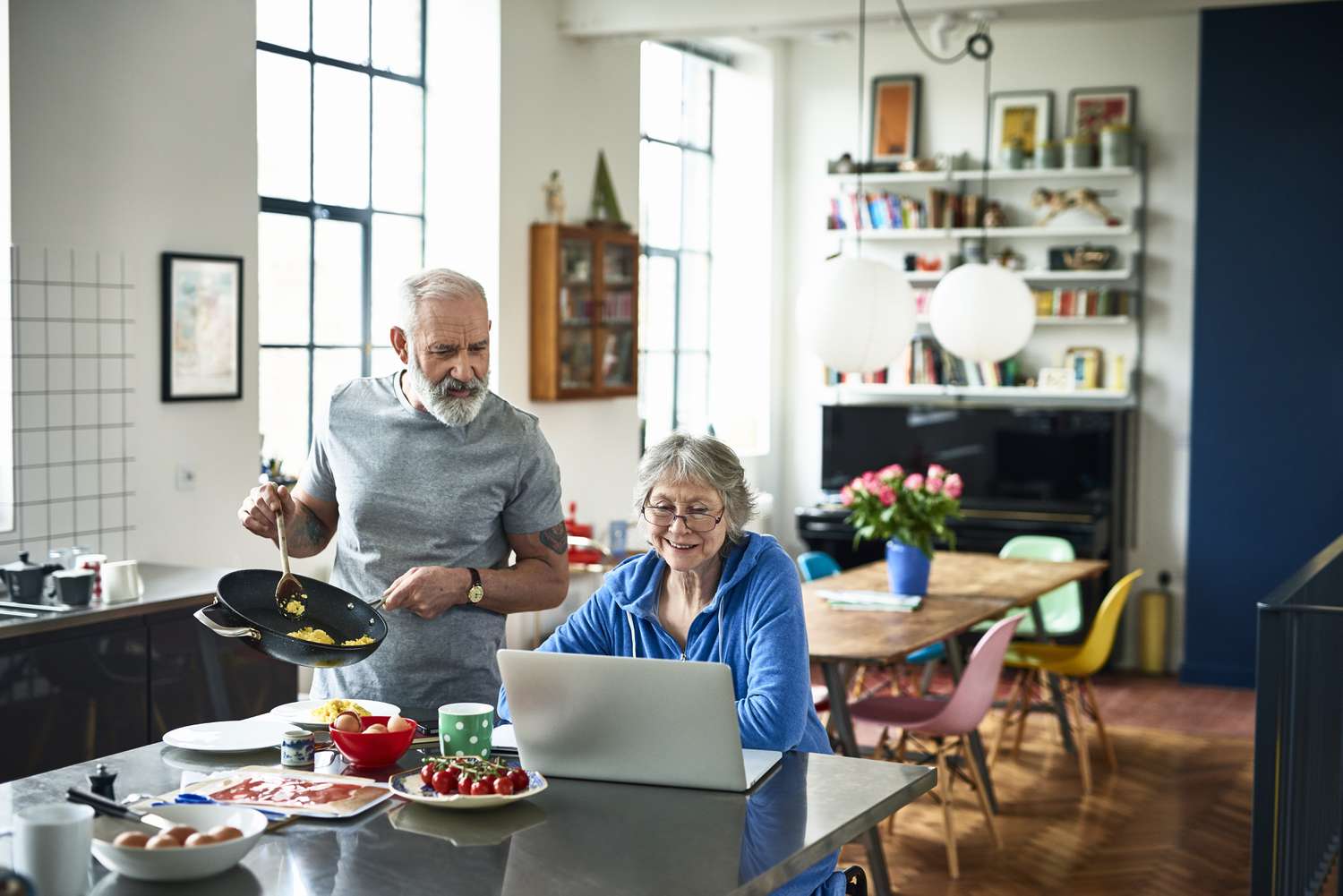 Retired couple in modern kitchen on the weekend, one surfing the web, the other holding a frying pan and serving scrambled eggs.