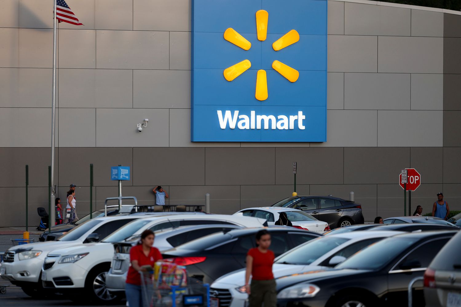 People and cars are seen outside a Walmart store, with the blue and yellow Walmart logo on the side of a gray building.