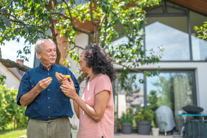 An older couple sharing an orange in the garden with their house in the background.