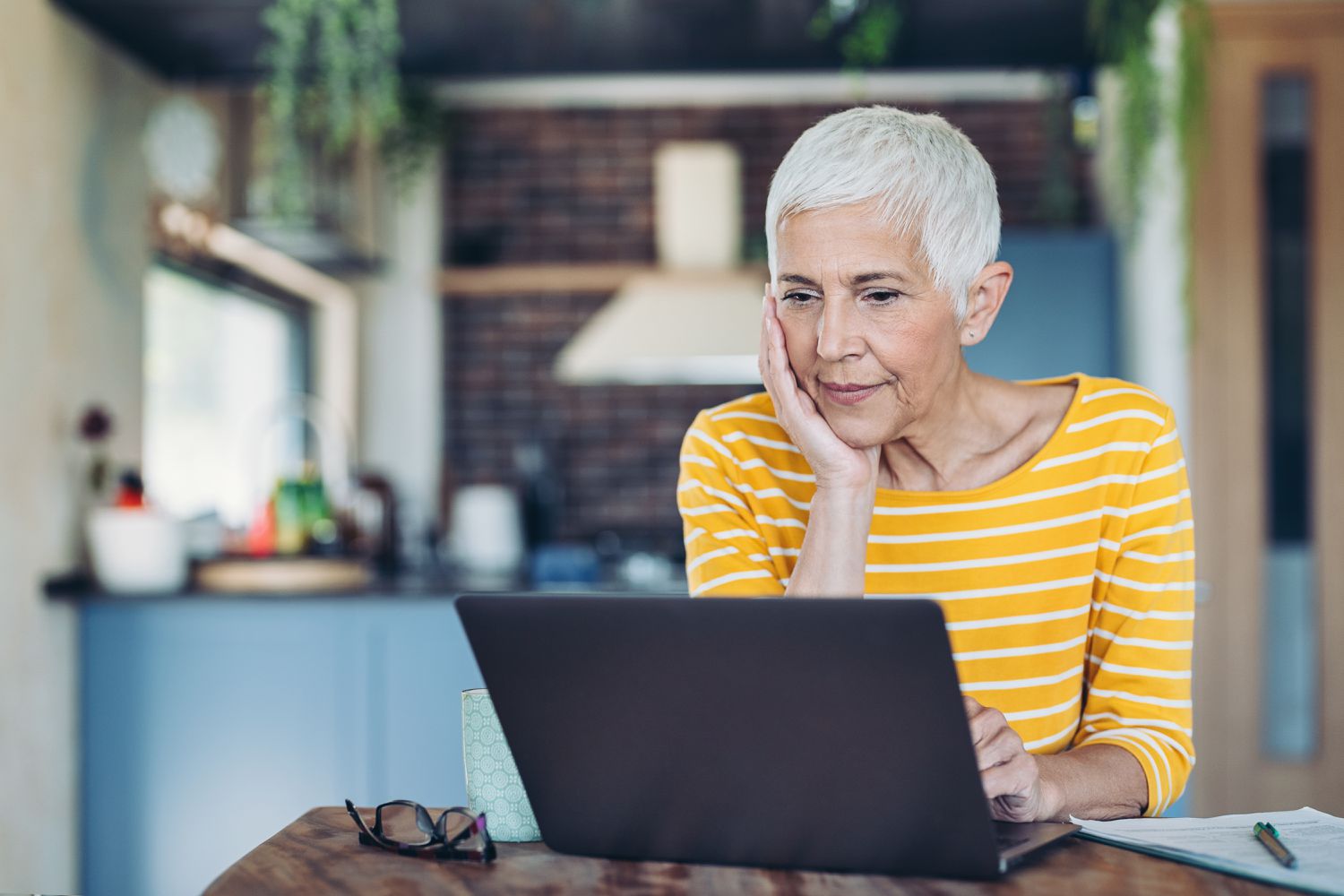 Woman working on a laptop in her kitchen, cupping her cheek in her right hand.