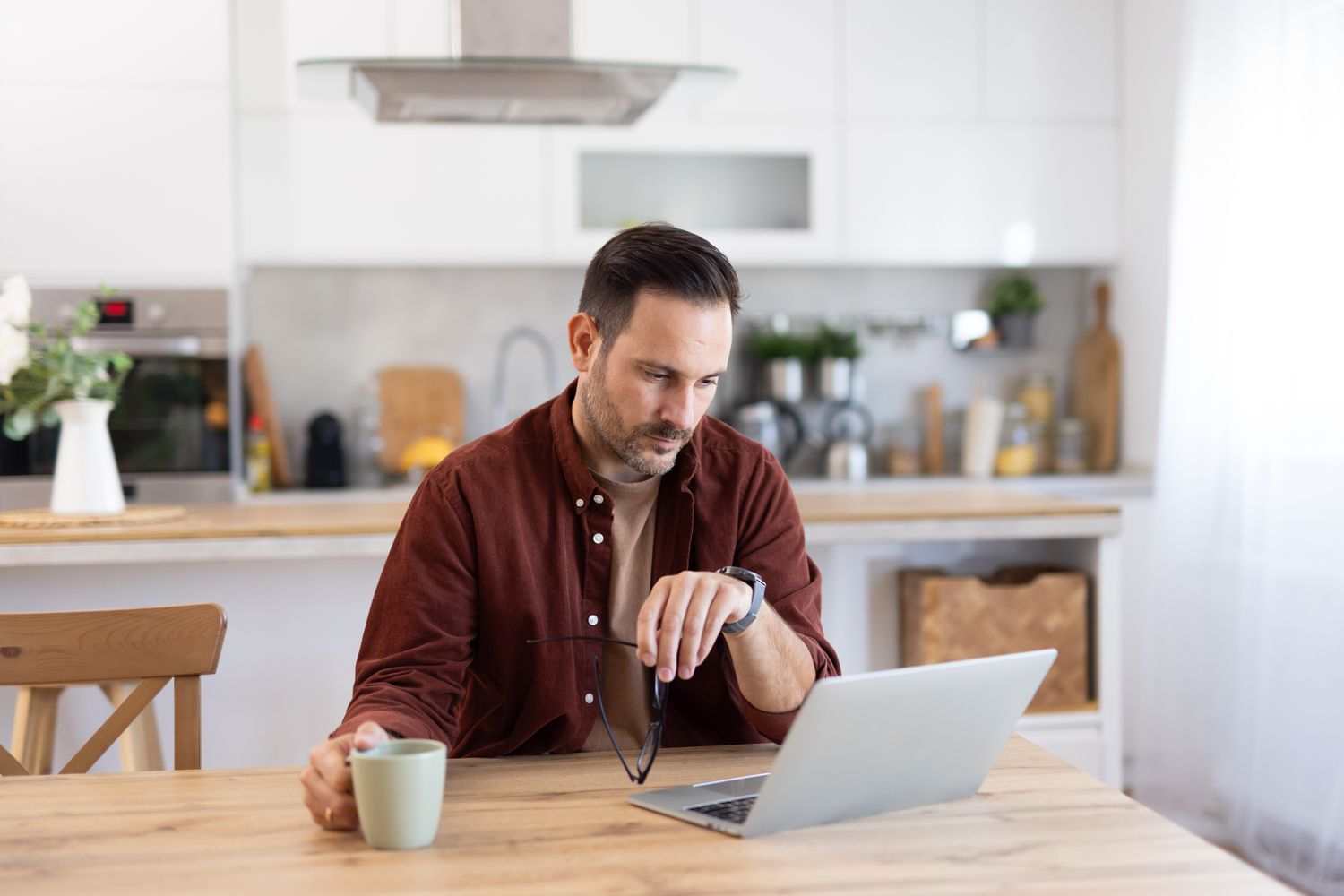 Man in his 30s sitting in his kitchen and looking seriously at news on his laptop