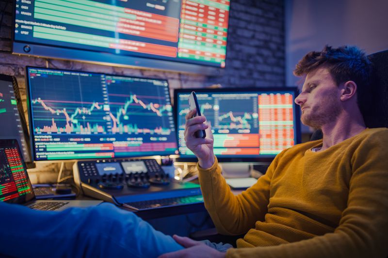 Trader looking at his phone in front of his desk with multiple monitors and programs.