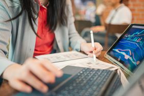 A woman writes on a piece of paper, while sitting in front of a laptop and a tablet displaying financial charts