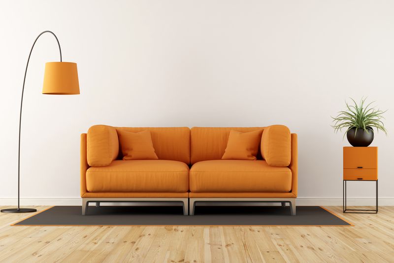 Orange sofa with cushions, rug, orange lamp, and orange side table against white wall.