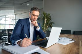 A man in a suit writing in a notebook in front of a laptop in an office. 