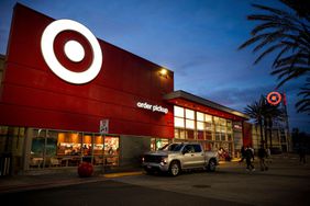 People and a silver truck are seen outside a Target store with a white Target logo on a red background, in front of a dark blue evening sky.
