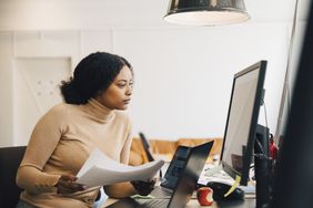 Focused female IT professional holding document while looking at computer in creative office