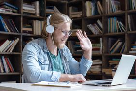 Happy student wear headphones waving hand looking at laptop computer screen video conference talking with online skype teacher communicating for distance education e learning in library